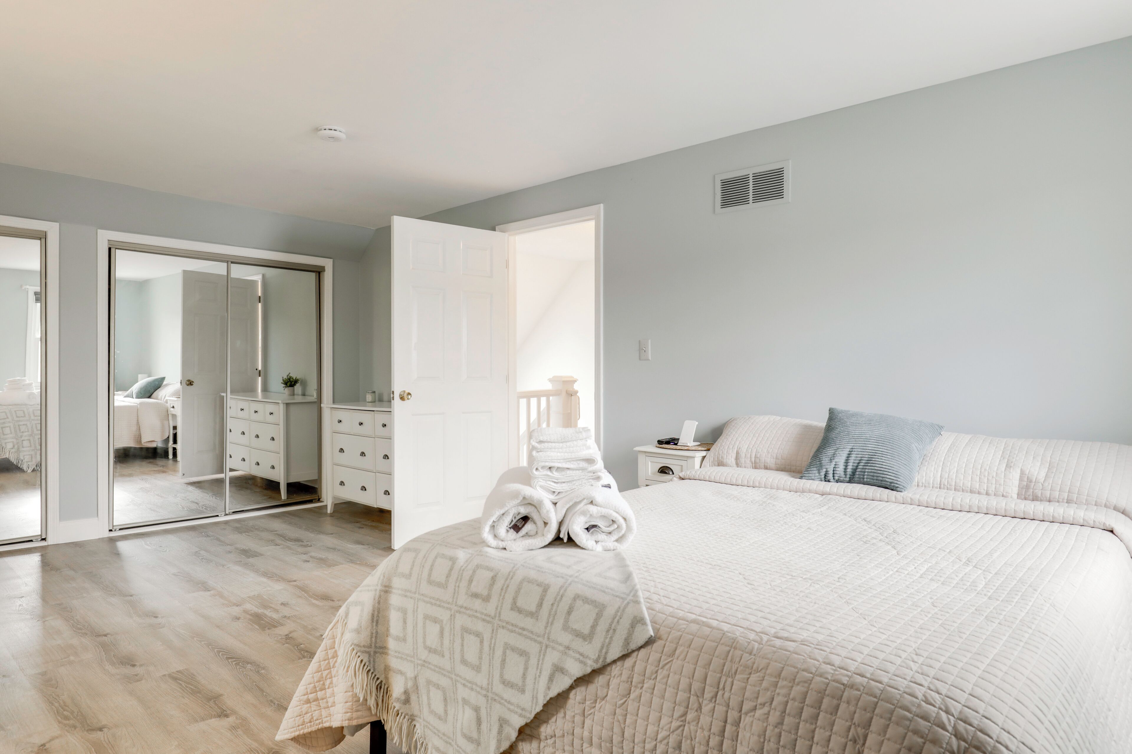 A bedroom with a bed, a bedside table, a nightstand, a dresser, a mirror, and a door leading to another room.