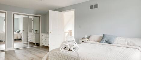 A bedroom with a bed, a bedside table, a nightstand, a dresser, a mirror, and a door leading to another room.