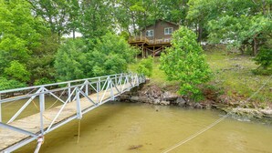 Un pont en bois enjambant une rivière aux eaux troubles, entouré d’une végétation luxuriante et d’une maison dotée d’une terrasse.