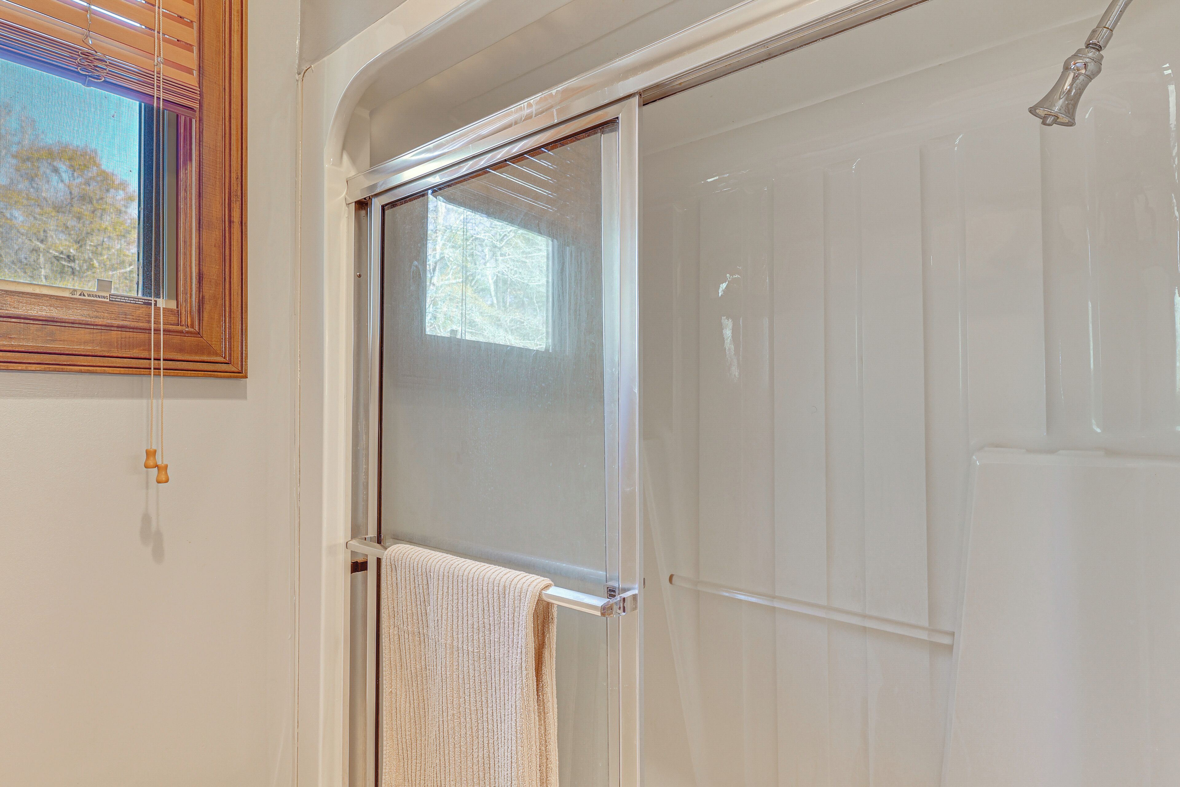 A bathroom with a glass shower door, a towel hanging on a rack, and a window with blinds.