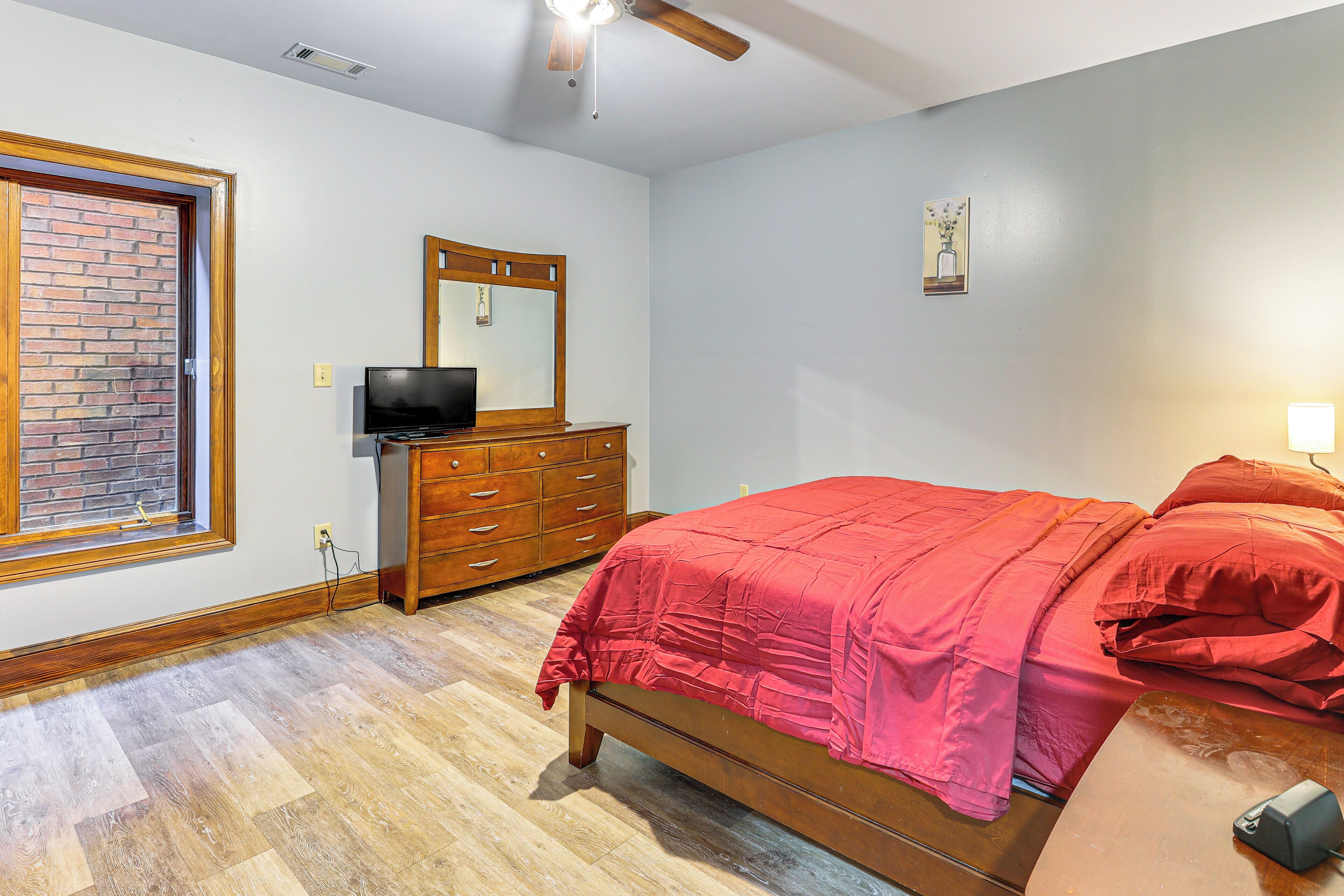 A bedroom with a wooden bed, a dresser, a television, and a window with a brick wall.