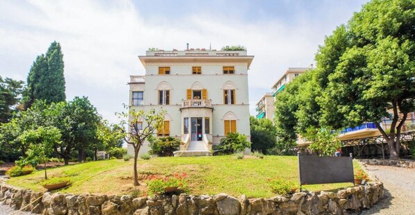 A two-story building with a central entrance, a stone wall surrounding the property, and a paved area in front.