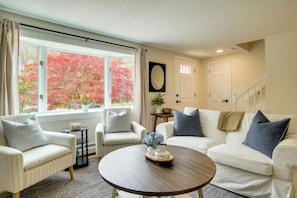 A living room with a sofa, armchair, and a coffee table. There is a window with a view of trees. - 3 Mi to Nauset Beach! Family Home w/ Private Yard (Orleans)