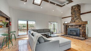 Un salon moderne doté d’une cheminée en pierre, d’un canapé d’angle gris, de planchers en bois et de grandes fenêtres offrant une vue sur l’extérieur.