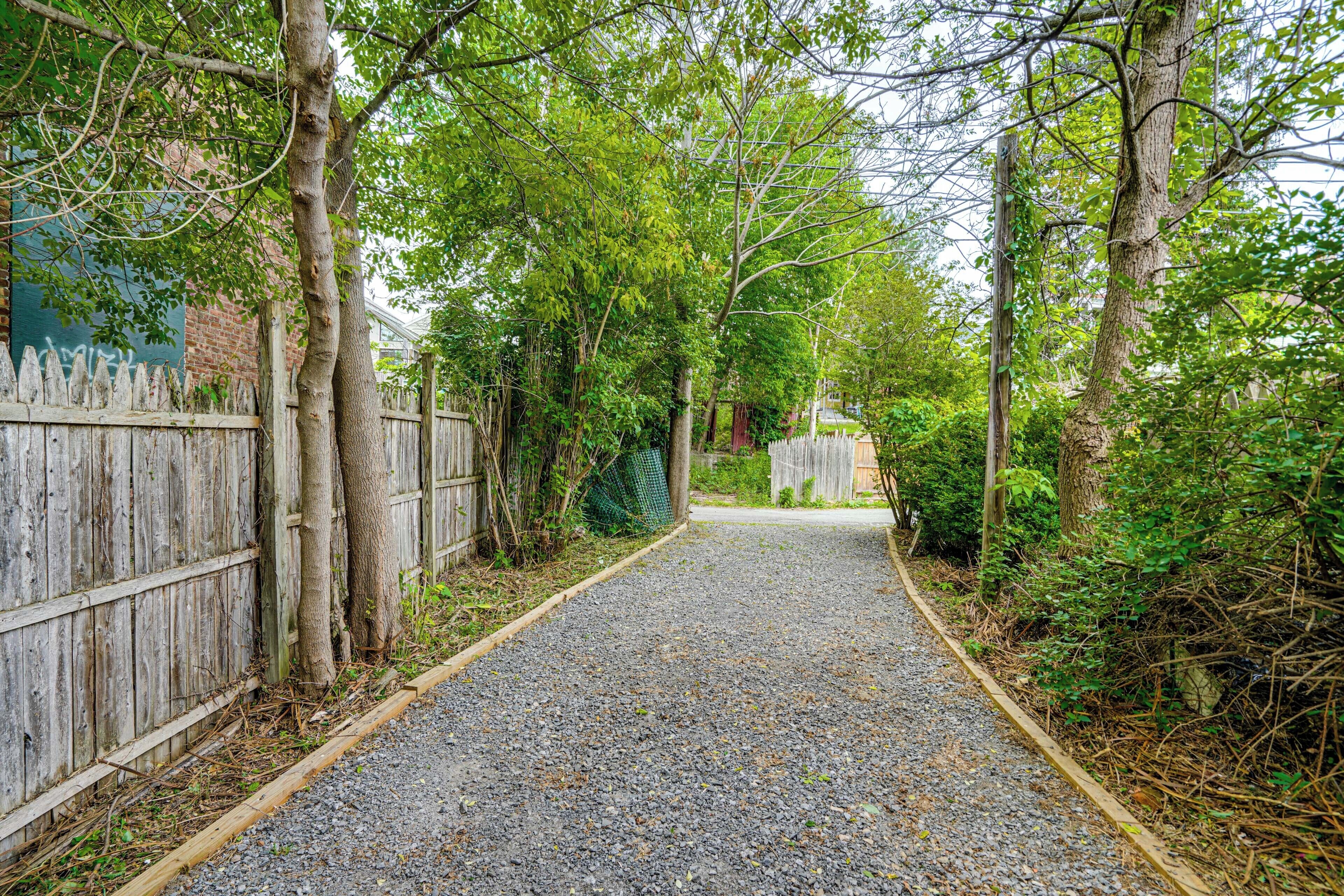 Un chemin de gravier bordé de clôtures en bois et d’arbres, qui mène à un bâtiment doté d’une porte verte.