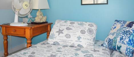 Een slaapkamer met een bed, nachtkastje met lamp en ventilator, een dressoir en een raam.