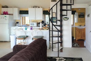 A compact kitchen with white cabinets, a microwave, and a coffee maker. There is a wooden chair and a brown sofa in the living area. A black spiral staircase leads to a bathroom with a sink and mirror. - Waterfront Studio w/ Dock & Beach Access in Truro! (Truro)
