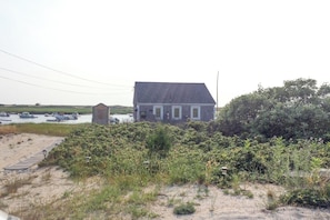 A house with a sloped roof, a small attached structure, and a sandy area with vegetation in the foreground. - Waterfront Studio w/ Dock & Beach Access in Truro! (Truro)
