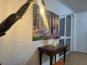 A room with a wooden floor, a painting of a forest, a table with a vase of flowers, and a hanging light fixture. - Vacation apartment Schwarzwaldblick Unterkirnach (Unterkirnach)