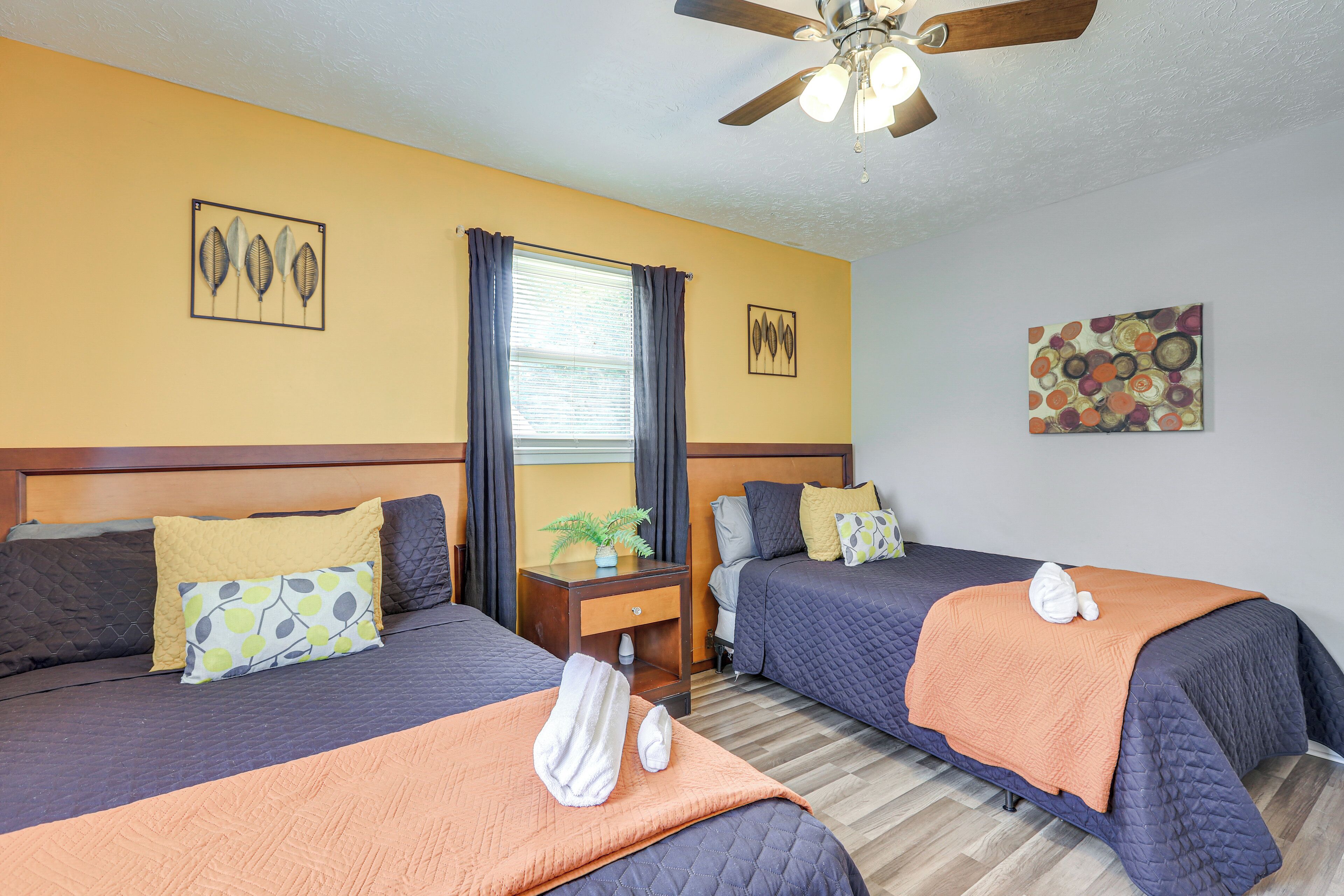 A hotel room with two beds, a ceiling fan, a window with curtains, and artwork on the walls.