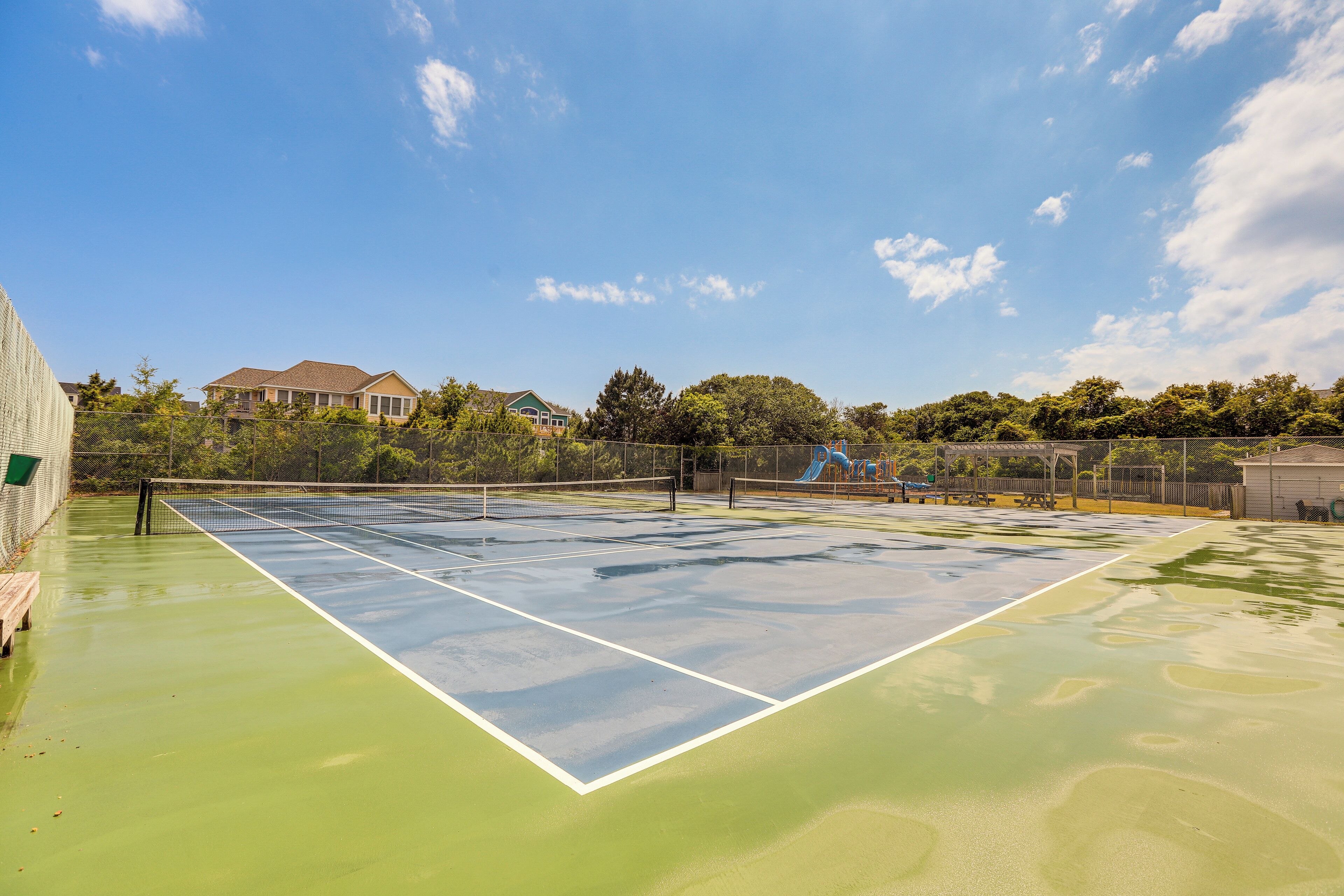 A tennis court with a net, surrounded by a fence, and houses in the background.