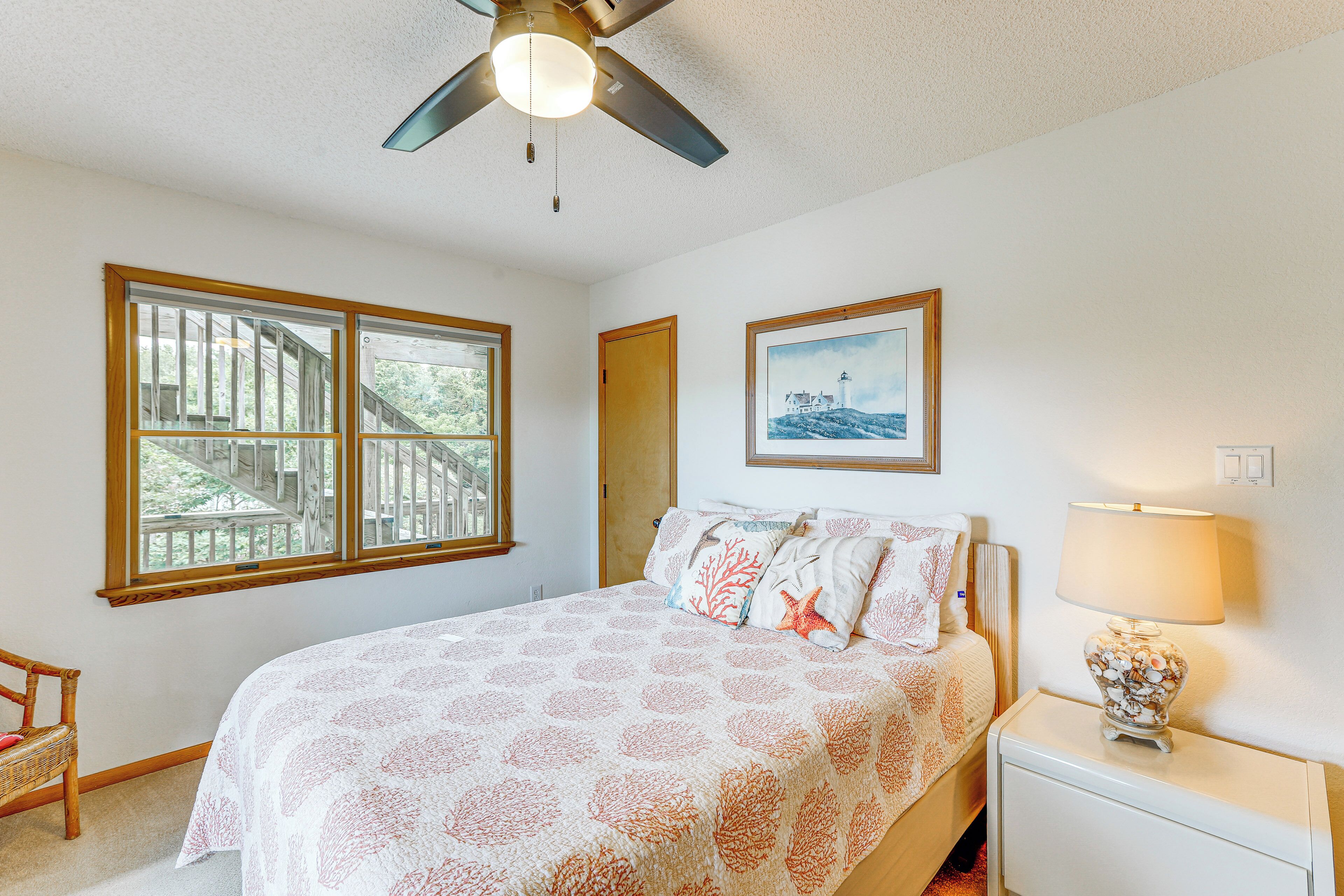Quarto com cama, mesa de cabeceira, luminária, cadeira, ventilador de teto e janela com vista para um deck de madeira.