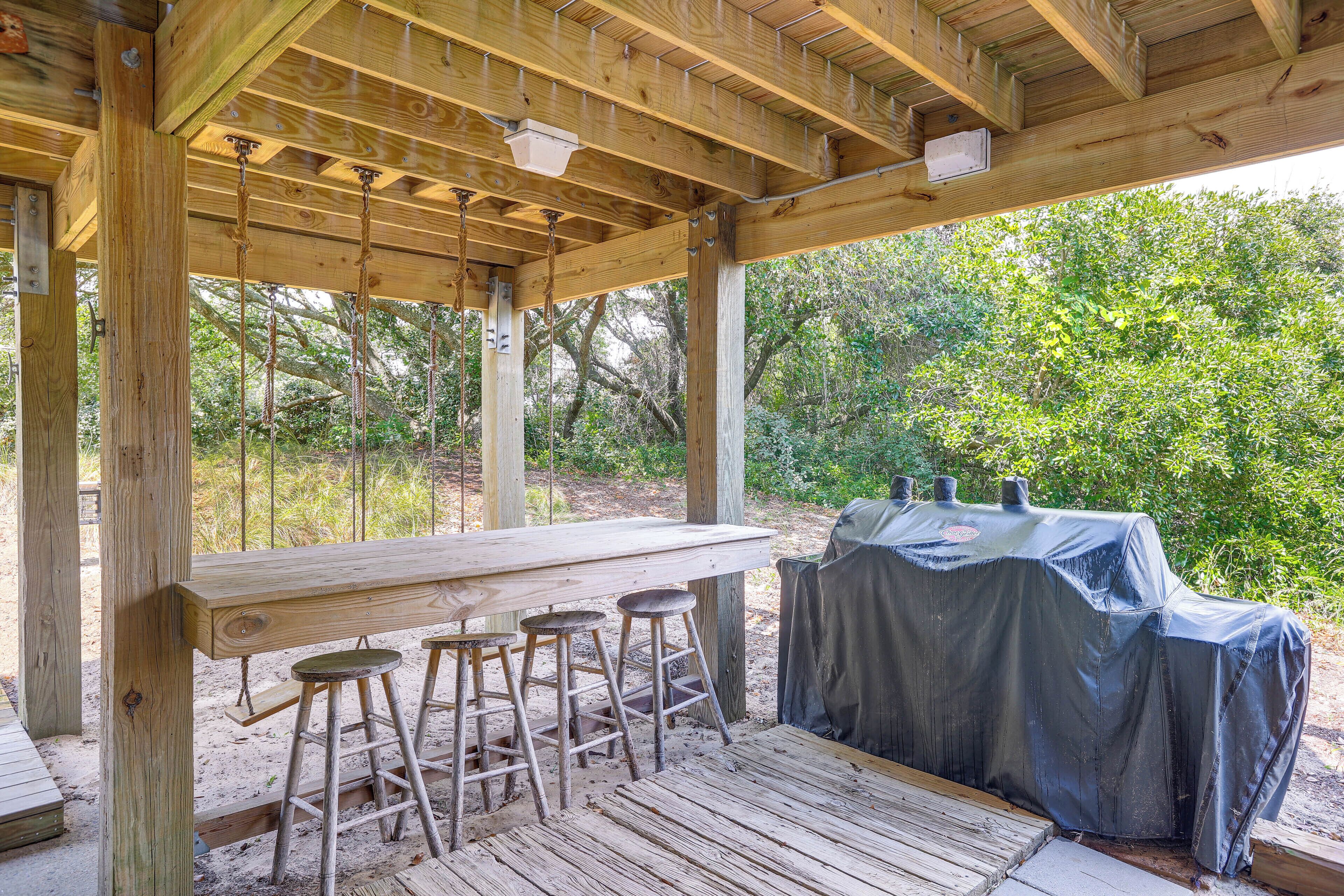 A wooden outdoor bar area with a long table and stools, a covered grill, and a view of trees.