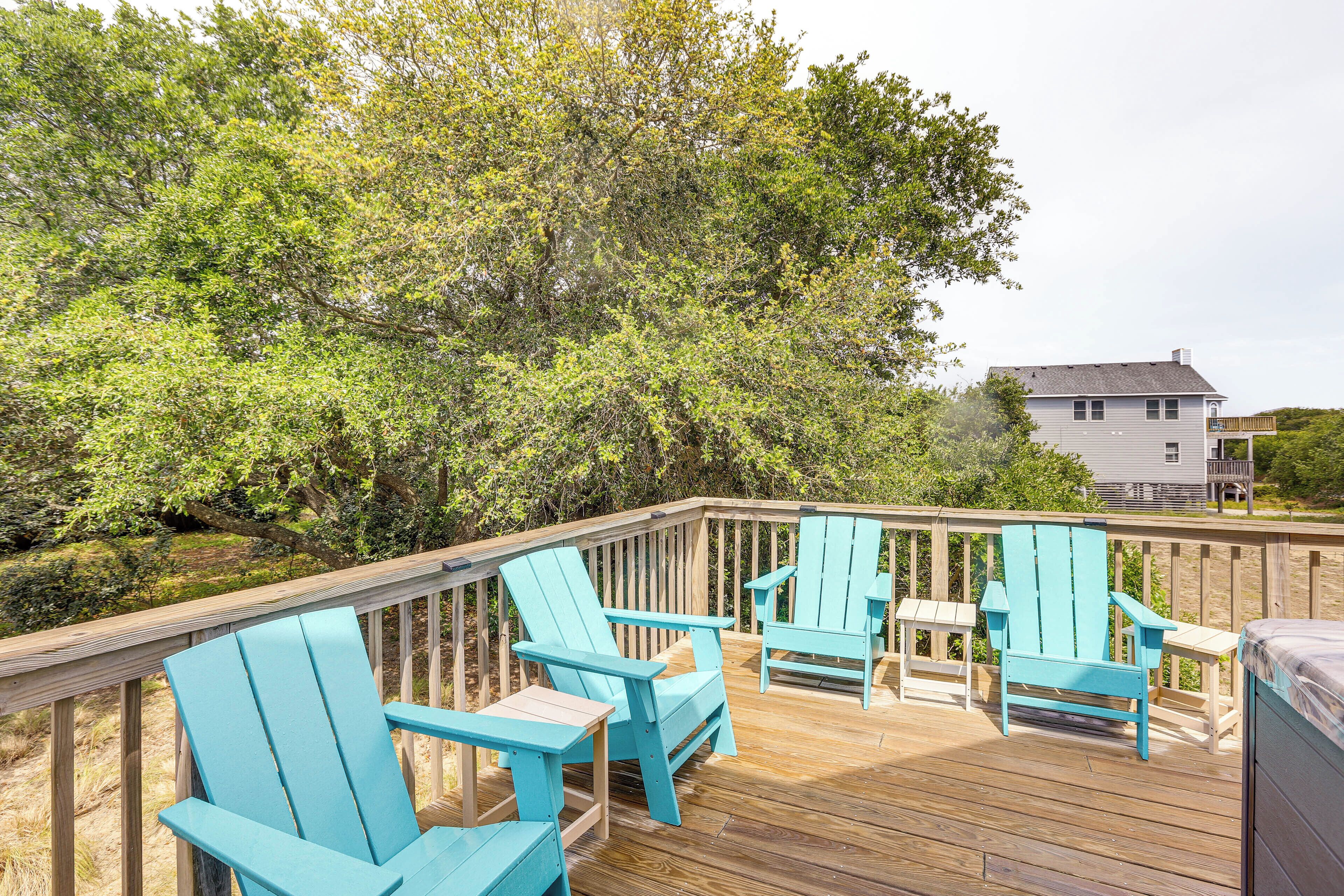 A wooden deck with blue Adirondack chairs and a small table, surrounded by lush greenery and a house in the background.
