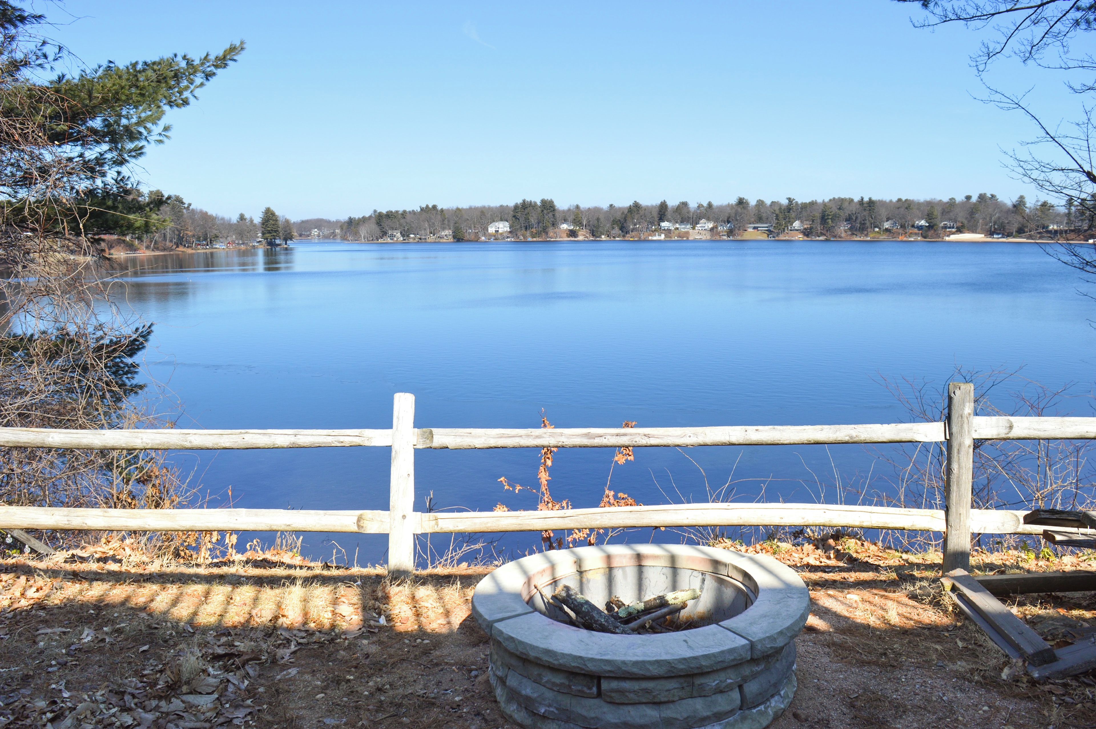 A calm lake with a wooden fence and a fire pit in the foreground.