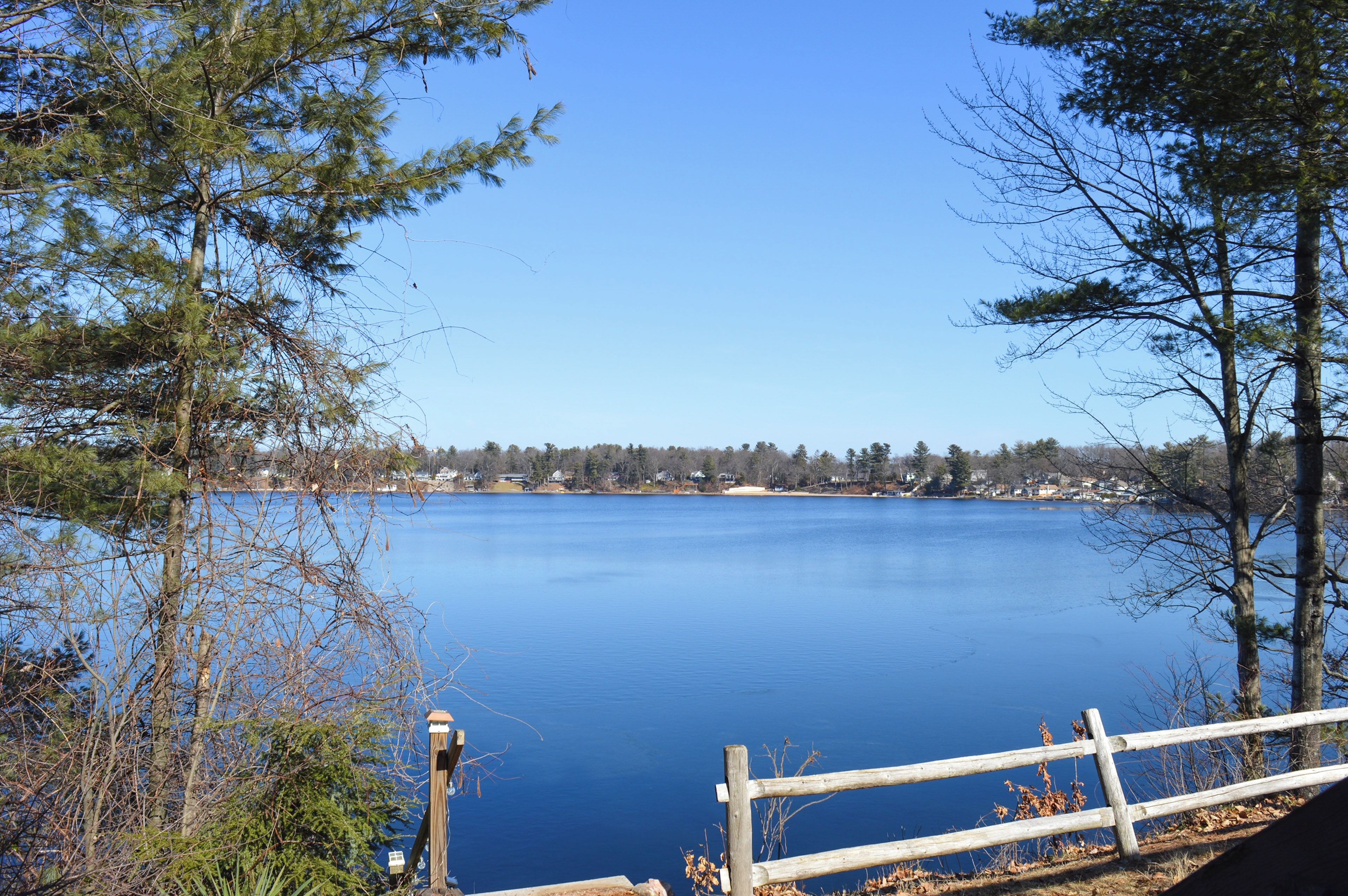 A calm lake with a wooden dock, surrounded by trees and houses in the background.