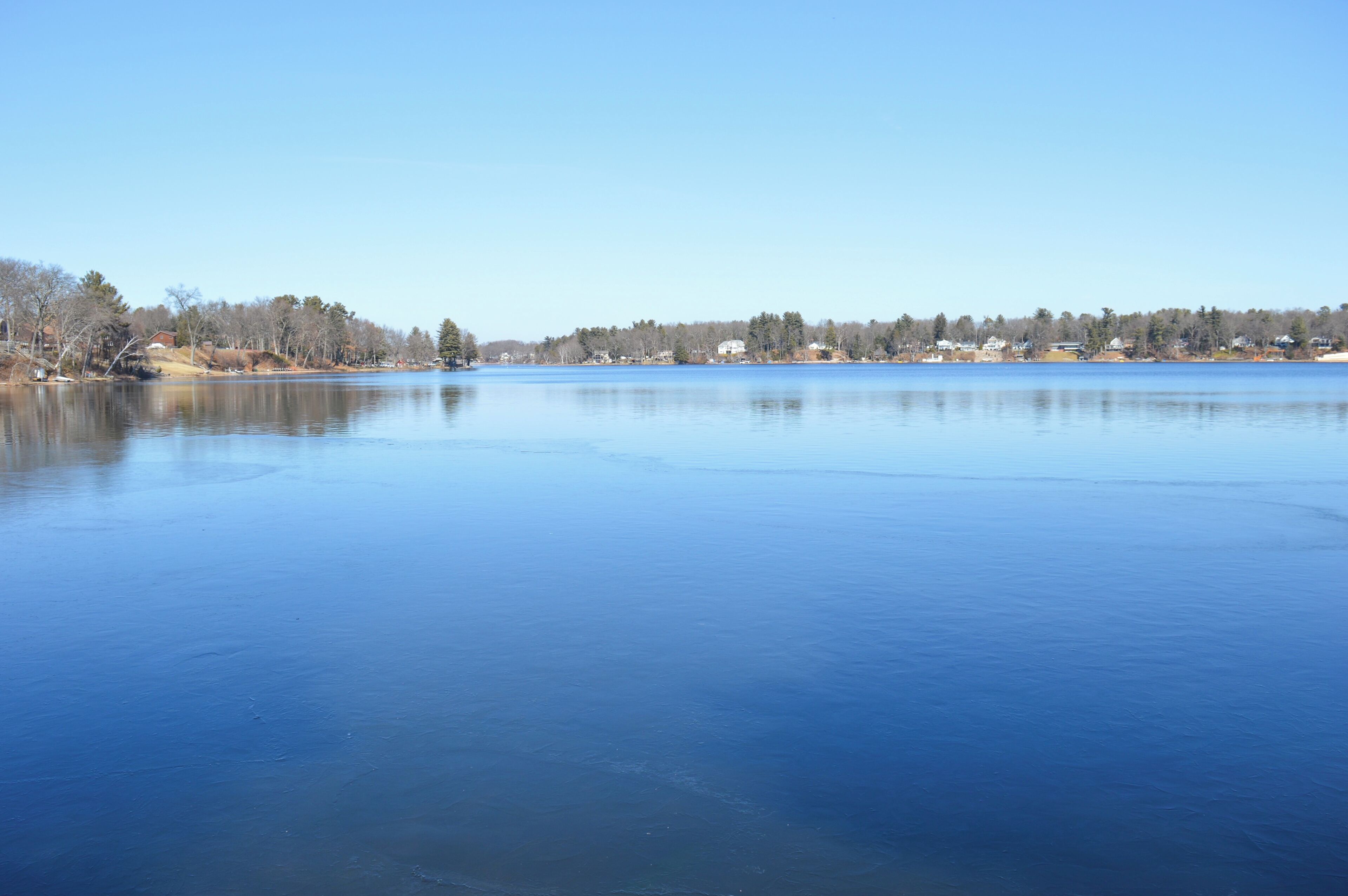 A calm lake with a clear sky, surrounded by trees and buildings.