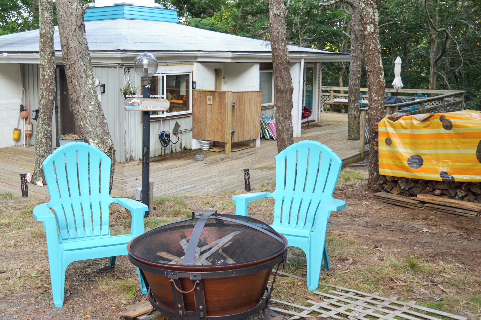 Een rustieke buitenruimte met twee turquoise Adirondack-stoelen, een metalen vuurkuil en een houten terras dat naar een huis met een blauw dak leidt.