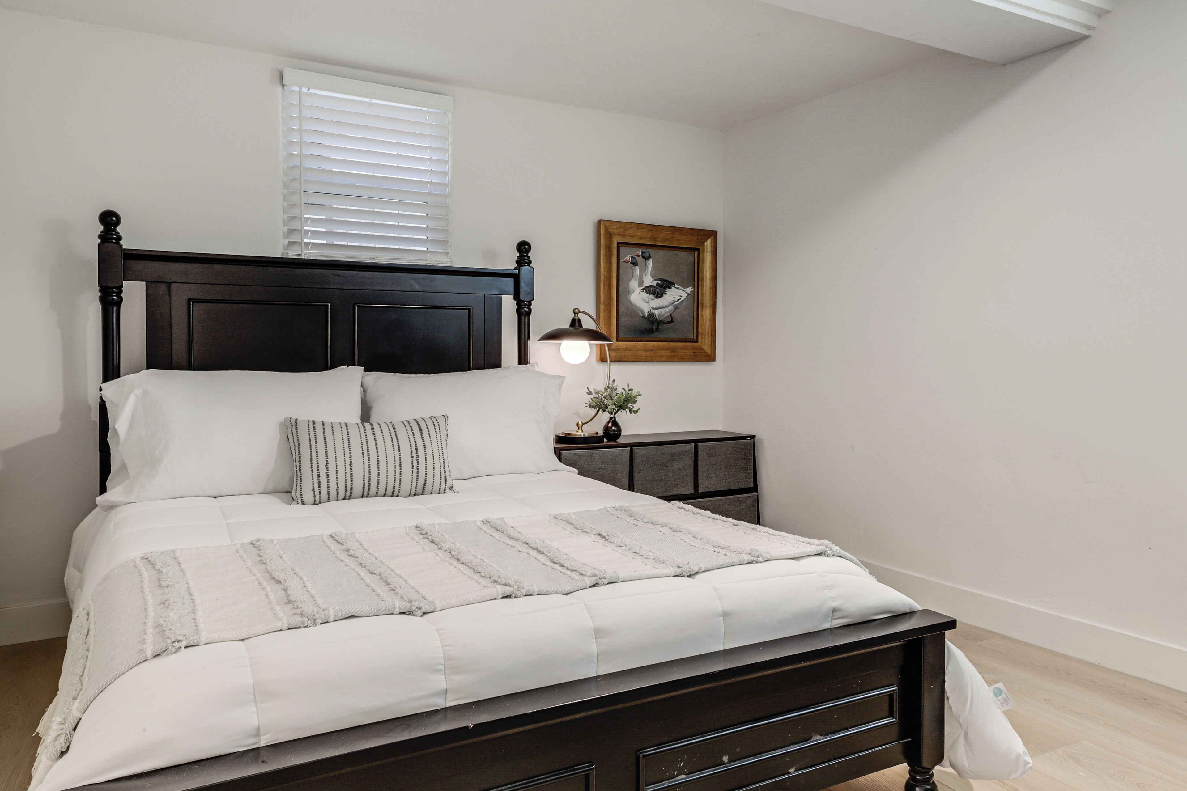 Een slaapkamer met een donker houten bed, witte beddengoed, een bedlampje, een ingelijst schilderij aan de muur en een raam met jaloezieën.