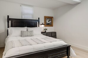 A bedroom with a dark wooden bed, white bedding, a bedside lamp, a framed picture on the wall, and a window with blinds.