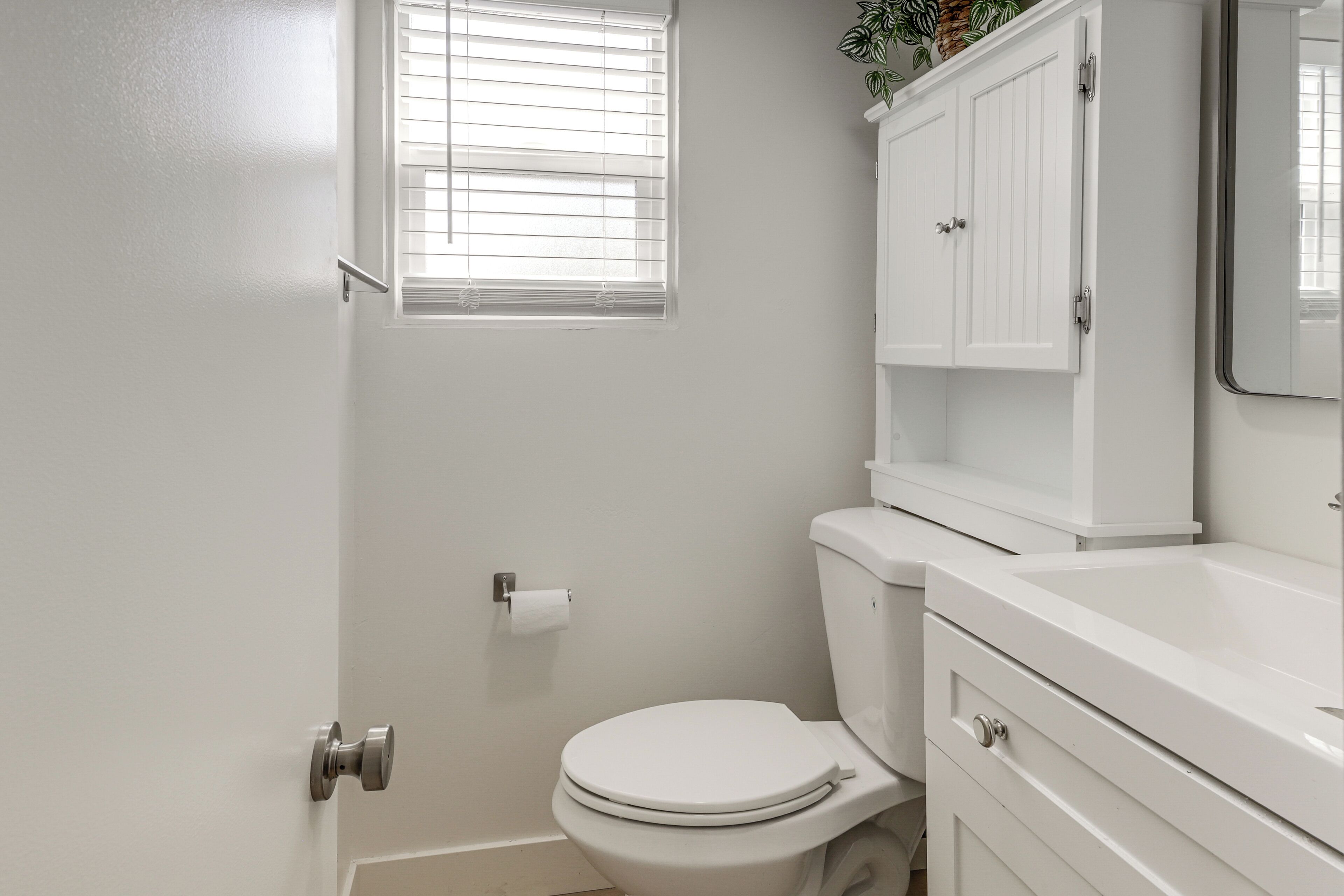 A bathroom with a toilet, a sink, a cabinet, and a window with blinds.