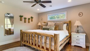 A bedroom with a wooden bed, a nightstand, a lamp, a mirror, and a window with a view of greenery.
