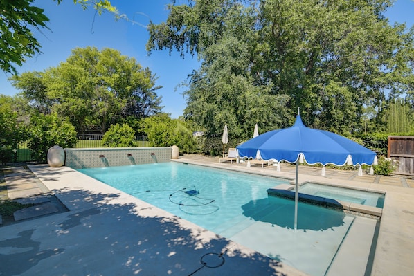 A rectangular swimming pool with a blue umbrella, surrounded by trees and a fence.