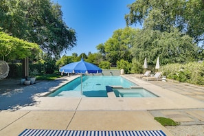 A rectangular swimming pool with a blue umbrella, surrounded by a stone deck, trees, and lounge chairs.