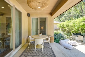 A covered patio with a round table and chairs, a woven ceiling light, and a view of a garden with outdoor furniture.