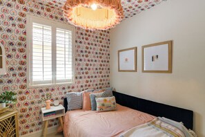 Une chambre avec un papier peint à motifs, un lit avec de nombreux coussins, une table de chevet avec une plante, et deux tableaux encadrés accrochés au mur.