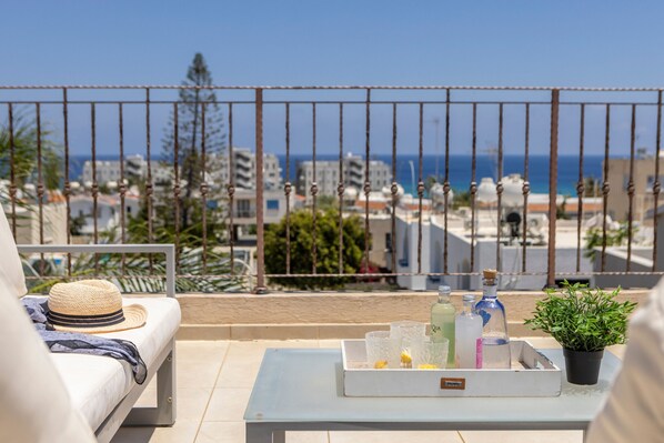 Ein Balkon mit einer weißen Couch, einem Hut, einem Tablett mit Getränken und einem Blumentopf.