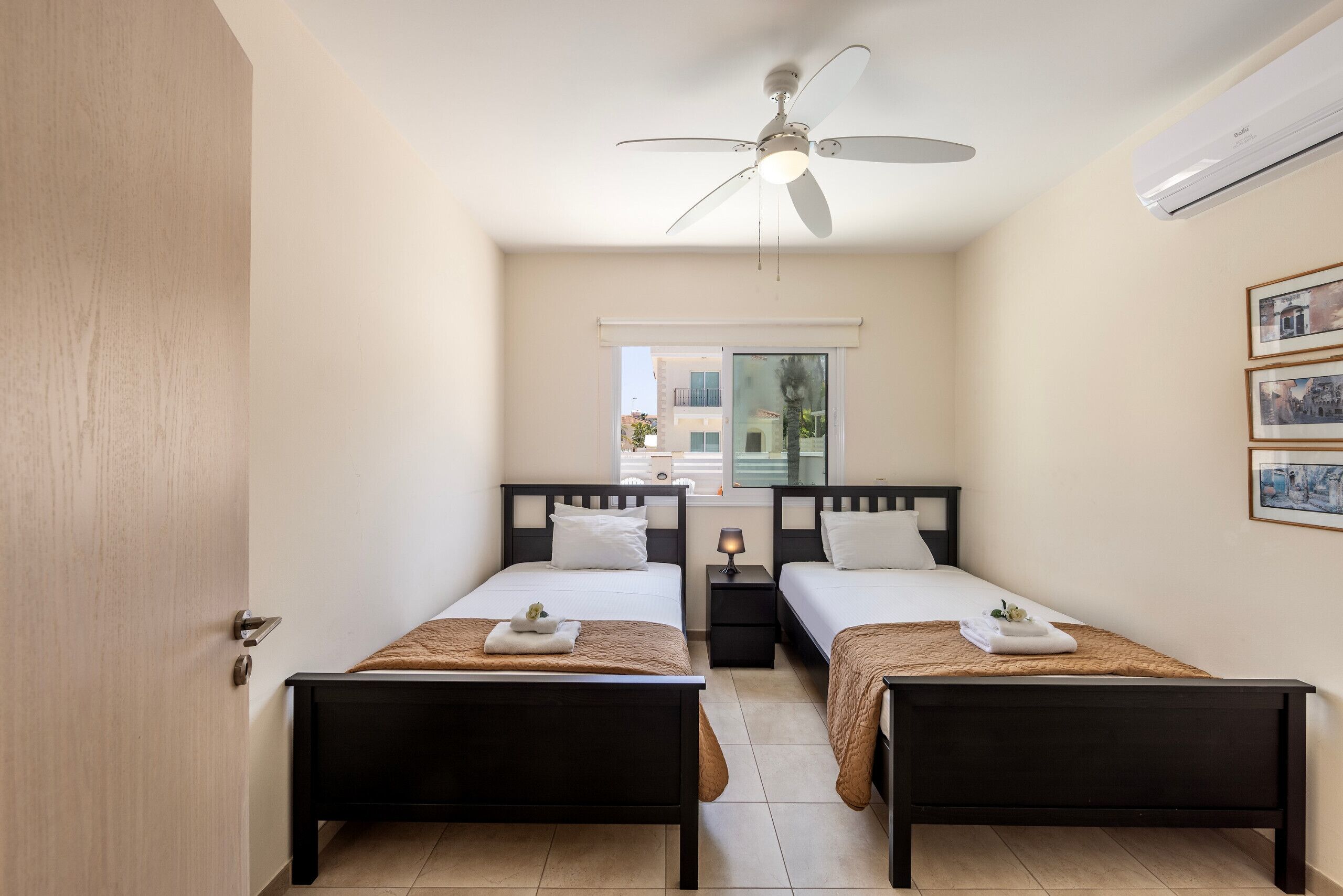 A hotel room with two beds, a ceiling fan, an air conditioner, a window with blinds, and framed pictures on the wall.