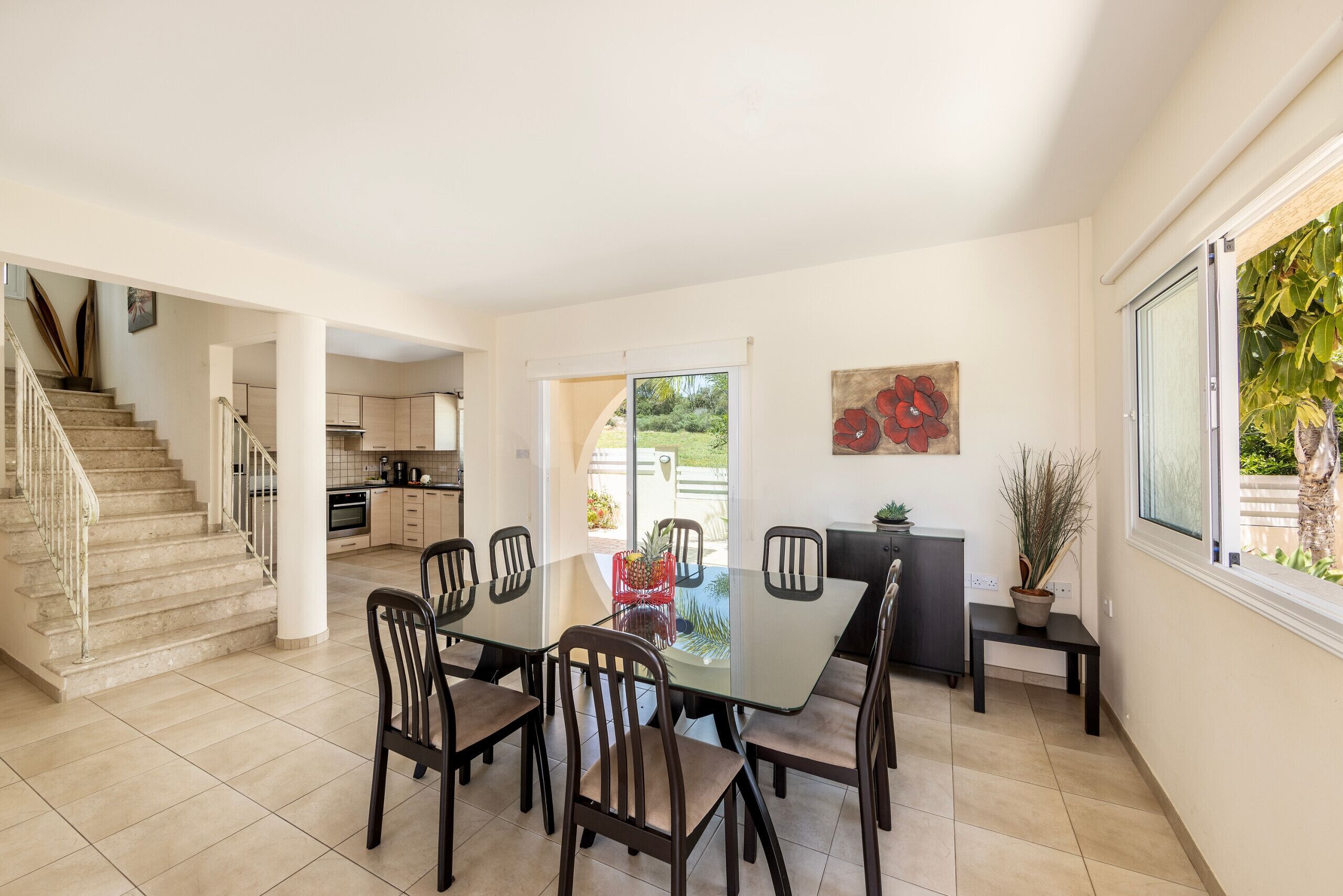 A spacious dining area with a glass table and chairs, a kitchen in the background, and a staircase leading to another level.