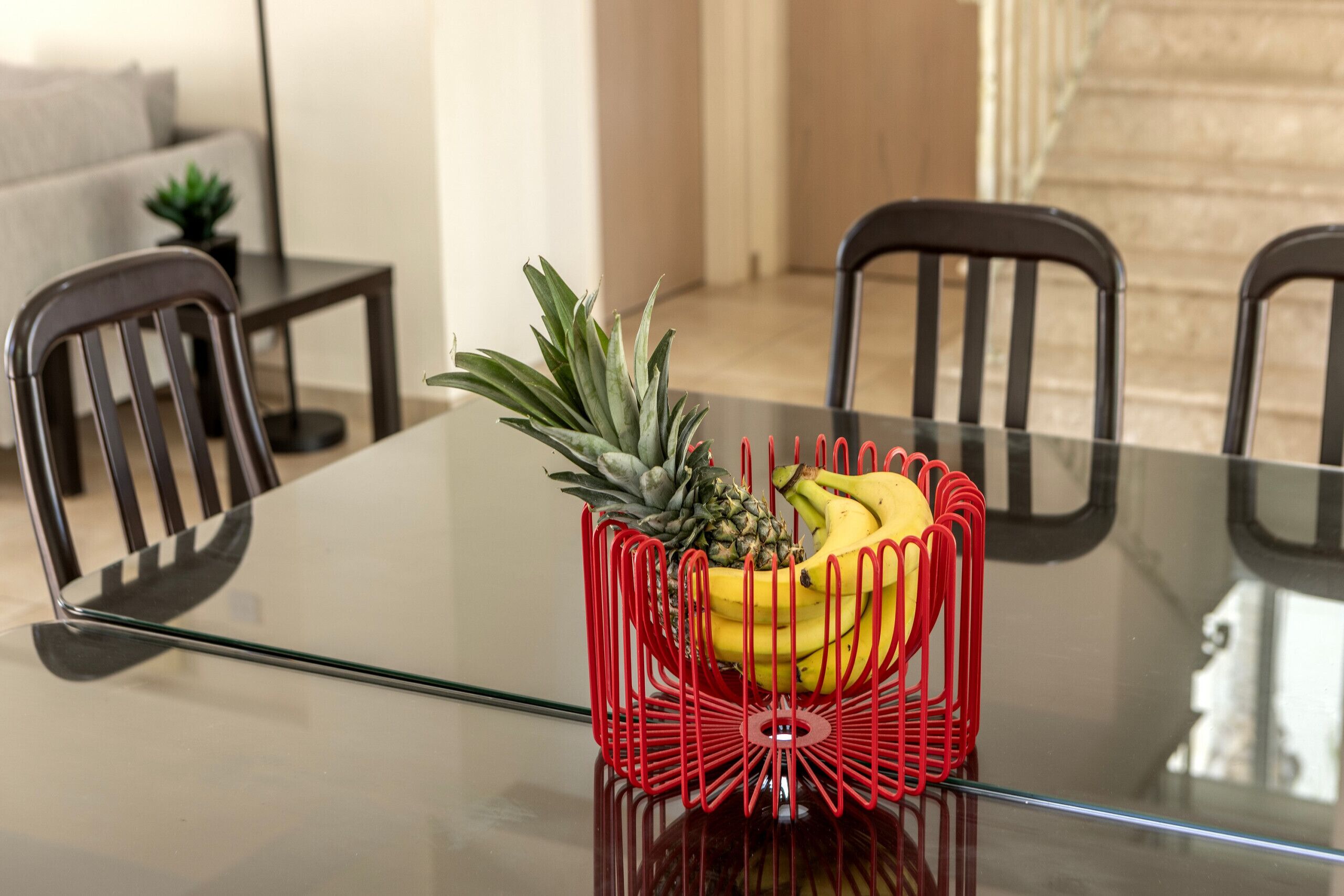 Un tavolo da pranzo con piano in vetro, circondato da sedie nere e un cesto di frutta rossa con banane e ananas.