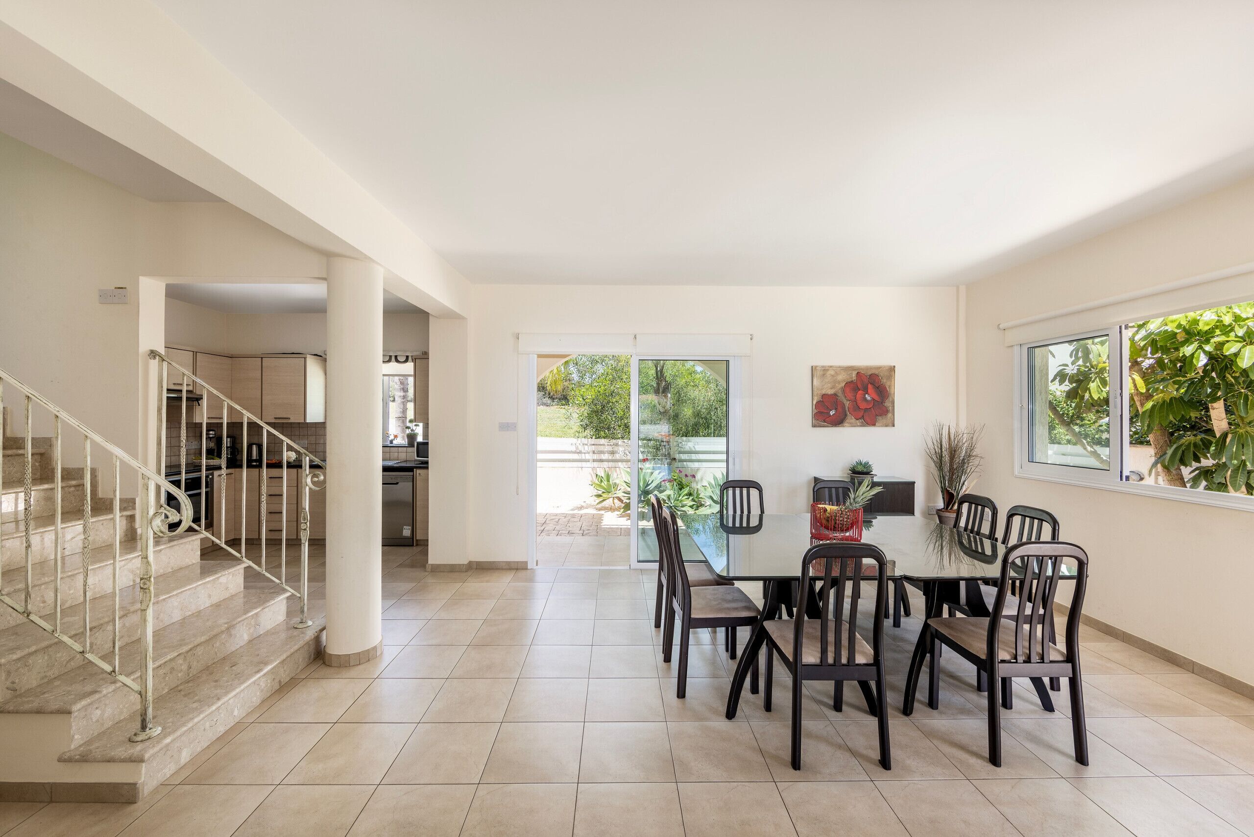 A spacious dining area with a glass table and chairs, a staircase, and a view of a garden through a sliding door.