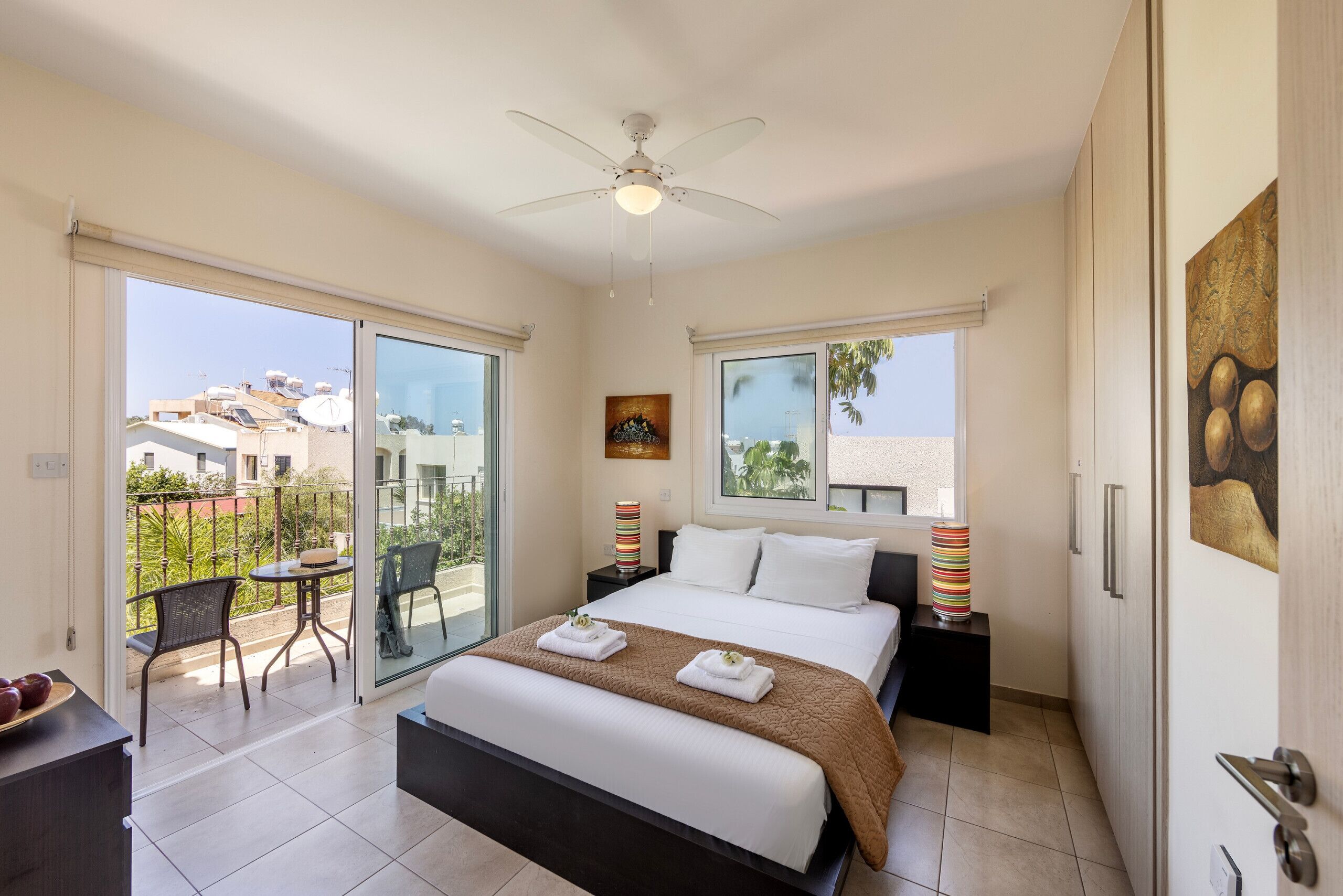 A bedroom with a bed, a ceiling fan, a balcony with a table and chairs, and a painting on the wall.