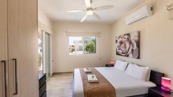 A hotel room with a bed, a ceiling fan, an air conditioner, a window with a view, and a painting on the wall.