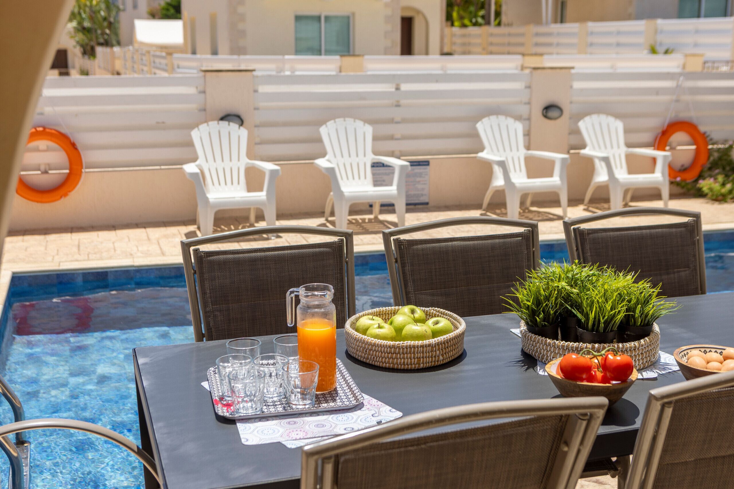 A poolside dining area with white chairs, a table with a pitcher of orange juice, and a bowl of apples and tomatoes.