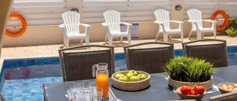 A poolside dining area with white chairs, a table with a pitcher of orange juice, and a bowl of apples and tomatoes.