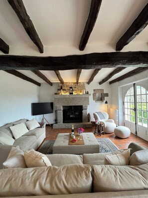 Uma sala de estar com lareira de pedra, vigas de madeira, um sofá, uma mesa de centro com uma garrafa e flores, e uma televisão.