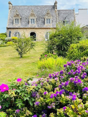 Um edifício de pedra com telhado inclinado, várias janelas e um jardim com flores roxas.