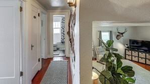 A hallway with a patterned rug, a wall-mounted light fixture, and a television in a living area. - Walkable Family Home in the Heart of Kalispell (Kalispell)