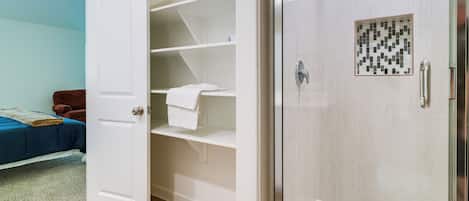 A bathroom with a glass-enclosed shower, a white door, and a blue bed in the adjacent room.