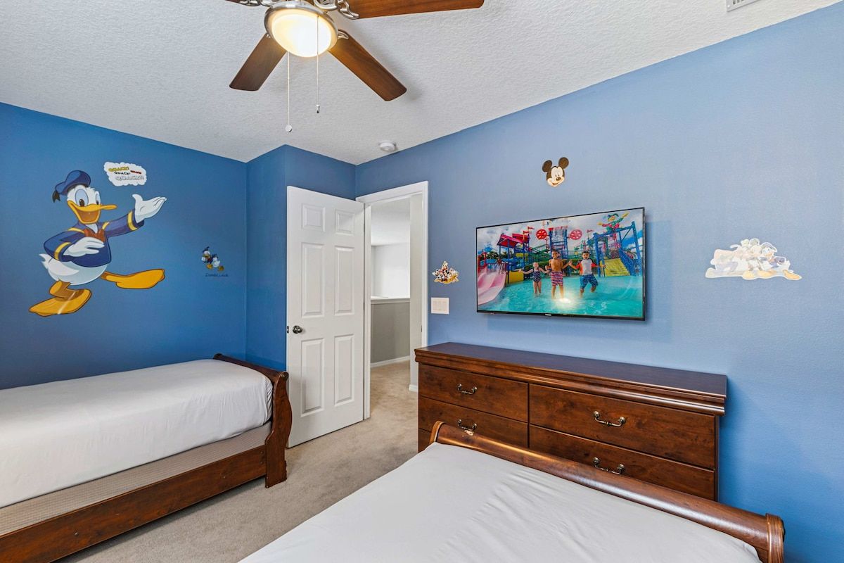 Une chambre avec deux lits simples, une commode en bois, un ventilateur de plafond et un mur décoré d’autocollants représentant des personnages de dessins animés.