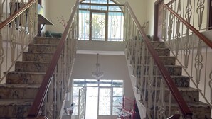 Uma escadaria imponente com degraus de mármore e grades de metal ricamente decoradas, que leva a um espaço amplo e iluminado, com uma grande janela e um candelabro.