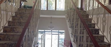 A grand staircase with marble steps and ornate metal railings, leading to a bright room with a large window and a chandelier.