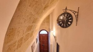 A narrow hallway with a clock on the wall, a wooden door at the end, and a carpeted floor. - B&B Luna Rossa (Altamura)