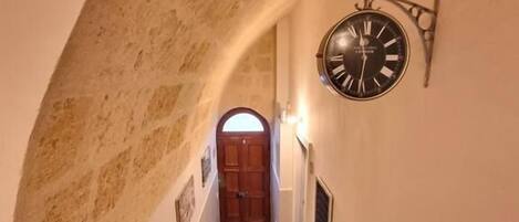 A narrow hallway with a clock on the wall, a wooden door at the end, and a carpeted floor.