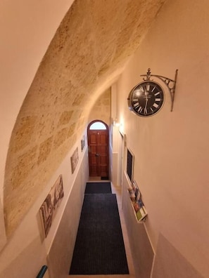 Un corridoio stretto con un orologio appeso al muro, una porta in legno in fondo e un pavimento rivestito di moquette.