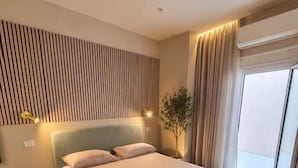 A modern hotel room with a large bed, a headboard with vertical lines, a bedside lamp, a small plant, and a window with curtains. - B&B Luna Rossa (Altamura)